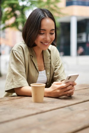 Foto de Retrato de una joven disfrutando de su café, bebiendo comida para llevar en el banco de la ciudad, usando un smartphone. Copiar espacio - Imagen libre de derechos