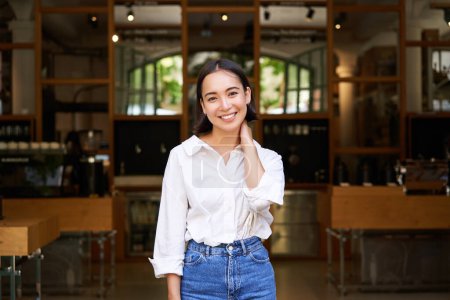 Foto de Retrato de mujer asiática joven linda y tímida, de pie frente a la entrada de la cafetería, tocando su cuello y sonriendo, presentando su negocio. - Imagen libre de derechos