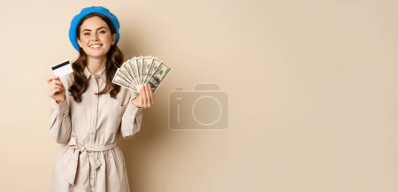 Foto de Microcredit and money concept. Young stylish woman showing credit card and dollars cash, smiling happy and satisfied, standing over beige background. - Imagen libre de derechos