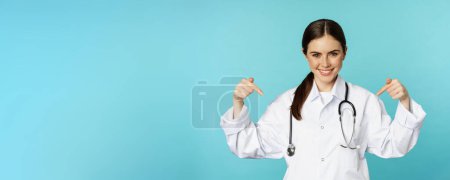 Photo for Smiling doctor medical worker, pointing fingers at logo, clinic banner, showing advertisement, wearing white coat, torquoise background. - Royalty Free Image