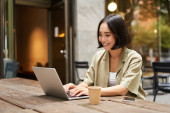 Young asian woman, digital nomad working remotely from a cafe, drinking coffee and using laptop, smiling. Poster #621611948