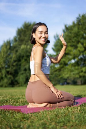 Téléchargez les photos : Portrait de fille asiatique en cours de fitness dans le parc, assis sur tapis de caoutchouc et main vague à la caméra, dire bonjour, méditation et yoga pratique. - en image libre de droit