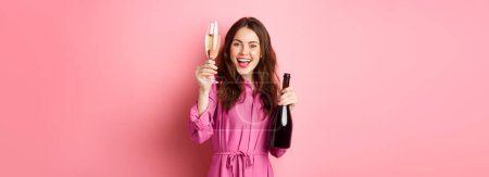 Celebration and holidays concept. Beautiful woman saying toast, raising glass of champagne and holding bottle, having fun at party, standing against pink background. Poster 621846908