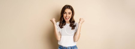 Photo for Enthusiastic woman winning and fist pump, celebrating victory, smiling excited, standing over beige background. - Royalty Free Image