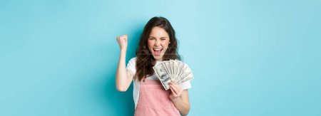 Glückliche junge Frau sieht aufgeregt aus, schreit vor Zufriedenheit und Triumph, gewinnt Geld, hält Dollarscheine in der Hand und macht eine Faustpumpe, steht vor blauem Hintergrund.