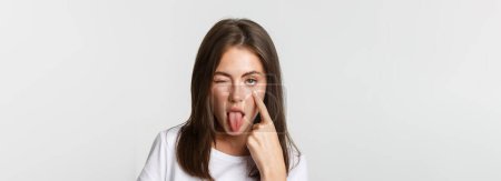 Foto de Primer plano de chica joven atractiva tonta mostrando la lengua y tirando del párpado, mirándote. - Imagen libre de derechos