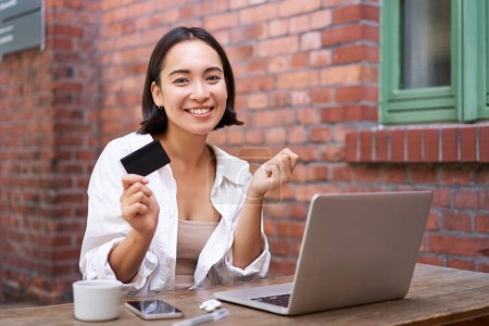 Photo for Beautiful young asian woman with credit card, sitting near laptop and smiling, paying bills, shops online, order smth on computer. - Royalty Free Image