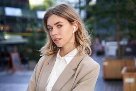 Photo for Confident businesswoman in headphones and beige suit, looking at camera with thoughtful gaze, posing outdoors. - Royalty Free Image