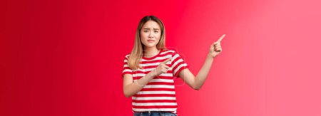 Photo for Whining displeased blond asian disappointed girl frowning hesitant grimacing unwilling come that place, pointing upper left corner reluctant, express apathy, upset bad choice, stand red background. - Royalty Free Image