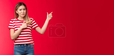 Téléchargez les photos : Mécontent lunatique asiatique copine agissant enfantin immature pointant coin supérieur droit bouleversé, fronçant les sourcils douteux insatisfait, regarder caméra hésitante et réticente, se tenir arrière-plan rouge non impressionné. - en image libre de droit