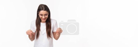 Photo for Waist-up portrait of enthusiastic, smiling happy brunette female student in glasses and t-shirt, looking pointing down, bottom advertisement, glancing pleased, standing white background. - Royalty Free Image