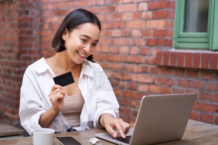 Foto de Mujer asiática sonriente sentada con computadora portátil, pagando con tarjeta de crédito por compras en línea, enviando los datos de su cuenta bancaria, sentada en la cafetería. - Imagen libre de derechos