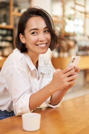 Téléchargez les photos : Plan vertical de la fille asiatique brune assise dans un café, à l'aide d'une application smartphone, bavarder sur un téléphone mobile et sourire. - en image libre de droit