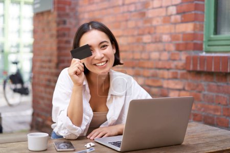 Photo for Happy young asian female, sitting with laptop and showing credit card, paying bills online, shopping in internet, sitting in cafe with coffee. - Royalty Free Image