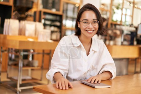 Téléchargez les photos : Jeune femme asiatique élégante, propriétaire d'entreprise dans des lunettes, assis dans un café avec ordinateur portable, souriant à la caméra. Concept de personnes et mode de vie - en image libre de droit