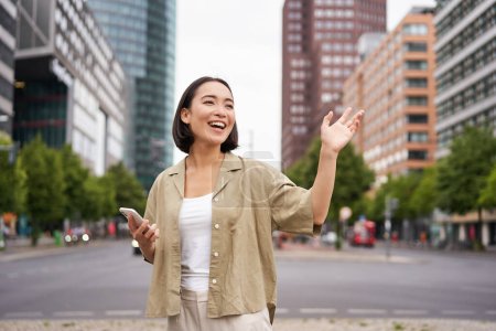 Téléchargez les photos : Heureuse fille asiatique passant par un ami et les saluant dans la rue, disant bonjour tout en marchant dans la ville, tenant smartphone. - en image libre de droit