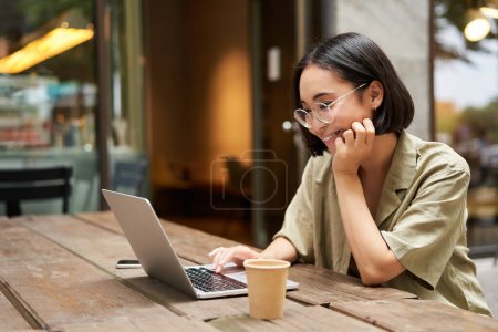 Photo for Portrait of smiling girl in glasses, sitting with laptop in outdoor cafe, drinking coffee and working remotely, studying online. - Royalty Free Image