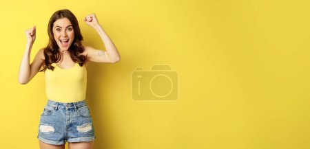 Photo for Enthusiastic young woman shouting and cheering, rejoicing, raising hands up and screaming in joy, chanting, standing over yellow background. - Royalty Free Image