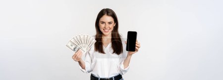 Foto de Woman showing mobile phone screen and cash, money, concept of microcredit and bank loans, standing over white background. - Imagen libre de derechos