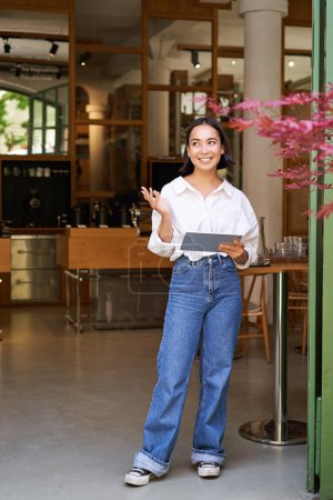 Foto de Tiro vertical de la sonriente mujer de negocios asiática, camarera de pie frente a la entrada de la cafetería, invitando a los huéspedes en el restaurante con tableta. - Imagen libre de derechos