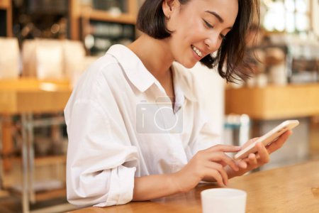 Téléchargez les photos : Portrait de jeune femme brune, assise avec du café et utilisant un smartphone dans un café, bavardant sur un téléphone portable. - en image libre de droit