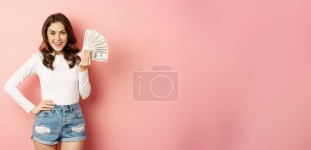 Foto de Attractive young woman holding money, cash in hands, concept of loans, microcredit and shopping, standing over pink background. Copy space - Imagen libre de derechos