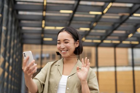 Téléchargez les photos : Heureux asiatique fille vagues à son smartphone caméra dit bonjour à ami sur vidéo chat, appeler quelqu'un, debout sur la rue. - en image libre de droit
