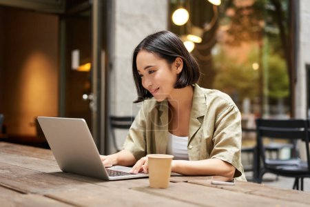 Photo for Young asian woman, digital nomad working remotely from a cafe, drinking coffee and using laptop, smiling. - Royalty Free Image