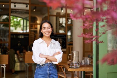 Foto de Retrato de una joven empresaria en su propia cafetería, gerente de pie cerca de la entrada y que invita, posando en camisa blanca lisa y jeans. - Imagen libre de derechos