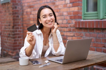 Enthusiastisch asiatisches Mädchen sitzt mit Kreditkarte und Laptop, bezahlt kontaktlos, kauft online per Computer ein.