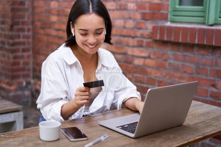 Photo for Smiling asian woman with laptop and wireless earphones, paying with credit card, buying online, sitting with cup of coffee. - Royalty Free Image