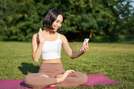 Téléchargez les photos : Portrait de fille asiatique dire bonjour sur smartphone, vague au téléphone mobile tout en faisant de la méditation, cours de yoga en ligne sur l'air frais dans le parc. - en image libre de droit
