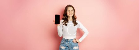 Téléchargez les photos : Femme sexy élégante montrant écran de téléphone mobile, interface d'application smartphone et souriant, boutique en ligne demonstarting, debout sur fond rose. - en image libre de droit
