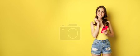 Technology and cellphone. Beautiful stylish girl order in online store, praying in application, using smartphone app and thinking, making decision, standing over yellow background.