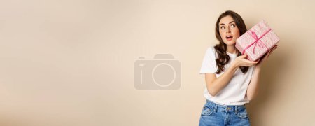 Foto de Celebration and holidays concept. Happy young girl looking intrigued, shaking box with gift, guessing whats inside wrapped present, standing over beige background. - Imagen libre de derechos