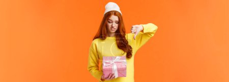 Photo for This gift sucks. Displeased problematic teenager, girl dont like present, looking at box with unsatisfied, displeased expression, grimacing skeptical, show thumb down in dislike, orange background. - Royalty Free Image