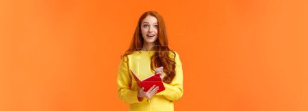 Photo for Inspired and upbeat, dreamy fascinated redhead girl writing down interesting lecture, smiling and gazing with admiration or enthusiasm, fill-in notebook schedule, standing orange background. - Royalty Free Image