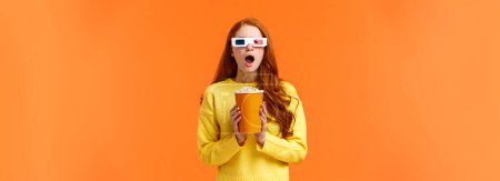 Photo for Shocked and tensed redhead woman seeing intense fight on screen as watching movie at cinema, wear 3d glasses, holding popcron, frowning and gasping concerned, orange background. - Royalty Free Image