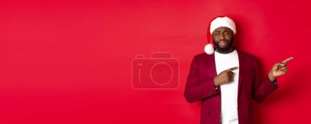 Photo for Christmas, party and holidays concept. Skeptical and unamused Black man looking with disdain, pointing fingers right at logo, standing in santa hat against red background. - Royalty Free Image