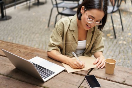 Foto de Retrato de una mujer joven que estudia en línea, sentado con el ordenador portátil, anotando, tomando notas y mirando la pantalla de la computadora, sentado en la cafetería al aire libre. - Imagen libre de derechos