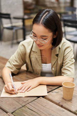 Foto de Tiro vertical de una joven asiática haciendo deberes, tomando notas, escribiendo algo, sentada en un café al aire libre y bebiendo café. - Imagen libre de derechos