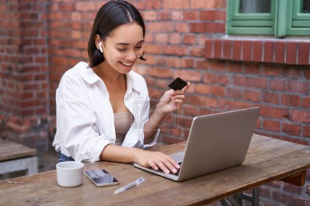 Lächelnde Asiatin mit Laptop und drahtlosem Kopfhörer, mit Kreditkarte bezahlen, online kaufen, mit Tasse Kaffee sitzen.