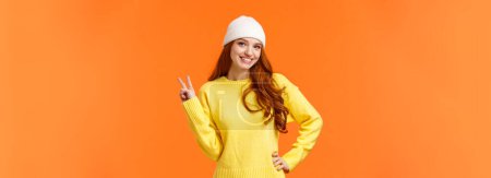 Téléchargez les photos : Moderne hipster fille jouer boules de neige avec des amis pendant les vacances d'hiver, montrant signe de paix comme posant photo près bonhomme de neige, portant bonnet blanc et pull sur fond orange
. - en image libre de droit