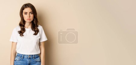 Photo for Sad and tired brunette woman looking drained and unamused, standing with pokerface against beige background. - Royalty Free Image