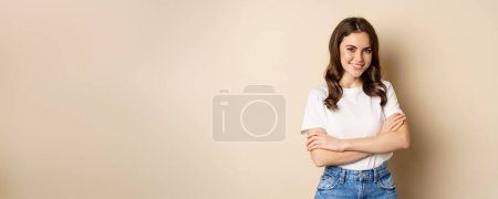 Foto de Retrato de una hermosa joven en camiseta blanca, sonriente y alegre, posando sobre fondo beige. - Imagen libre de derechos
