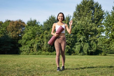 Téléchargez les photos : Femme sportive élégante avec tapis en caoutchouc pour le yoga, dit bonjour, vagues à vous, prêt pour l'entraînement en plein air dans le parc sur la pelouse. - en image libre de droit