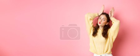 Foto de Chica linda y tonta mostrando conejito de Pascua o orejas de conejo por encima de la cabeza, labios pucker, haciendo cara de beso, de pie sobre fondo rosa. - Imagen libre de derechos