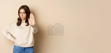 Foto de Stop. Serious and confident woman showing extended arm palm, prohibit, forbid smth, blocking something, standing over beige background. - Imagen libre de derechos