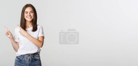 Téléchargez les photos : Satisfait fille brune souriante pointant des doigts coin supérieur gauche, montrant le logo. - en image libre de droit