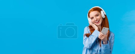 Photo for Lovely young dreamy girl fantasizing as listening favorite songs, voice of singer, tilt head and touching headphone as enjoying music earphones, holding smartphone and smiling, blue background. - Royalty Free Image
